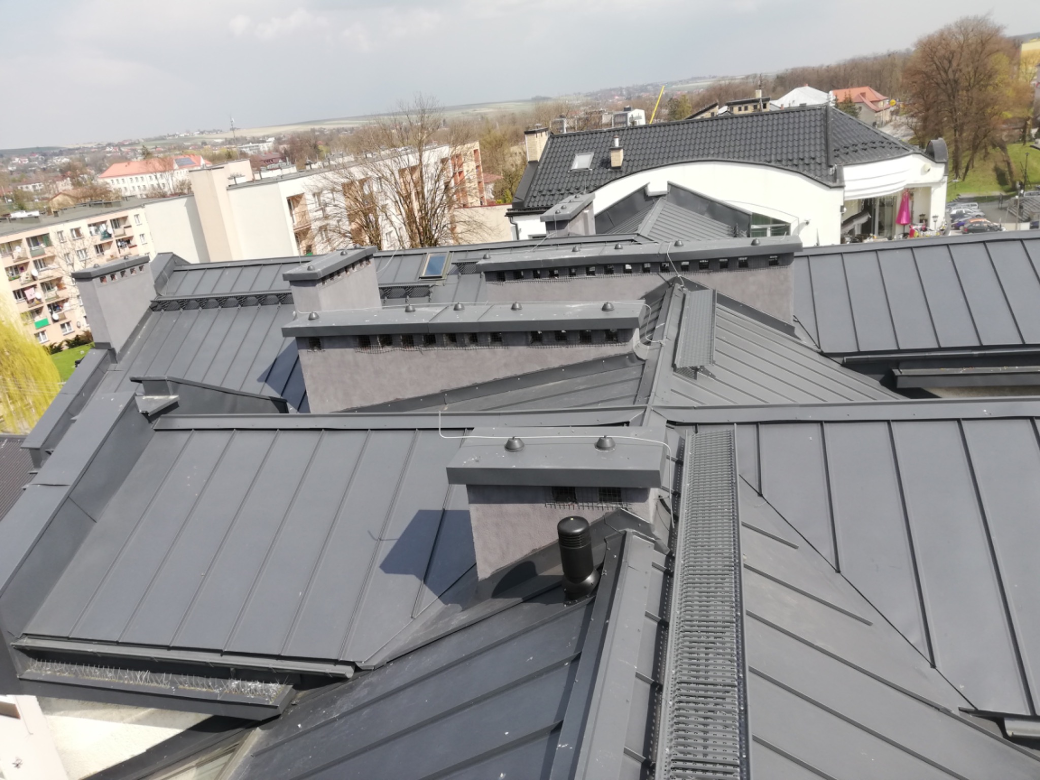 Szary metalowy dach z kominami i zabezpieczeniami przed ptakami, widok z góry na tle zabudowy miejskiej.