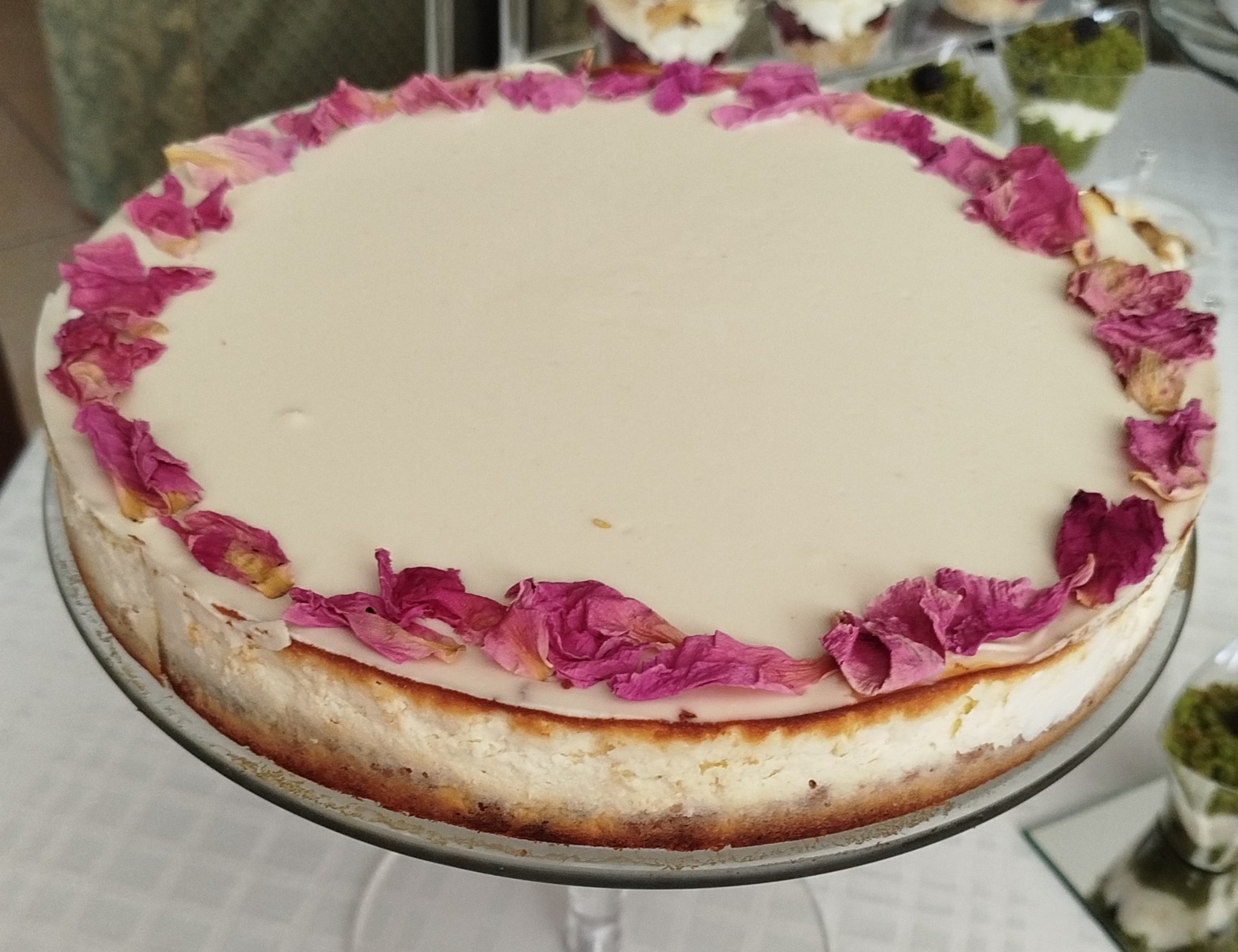 Okrągły sernik na szklanej paterze, udekorowany brzegiem z płatków róż w odcieniach różu i fioletu, z deserami w tle.