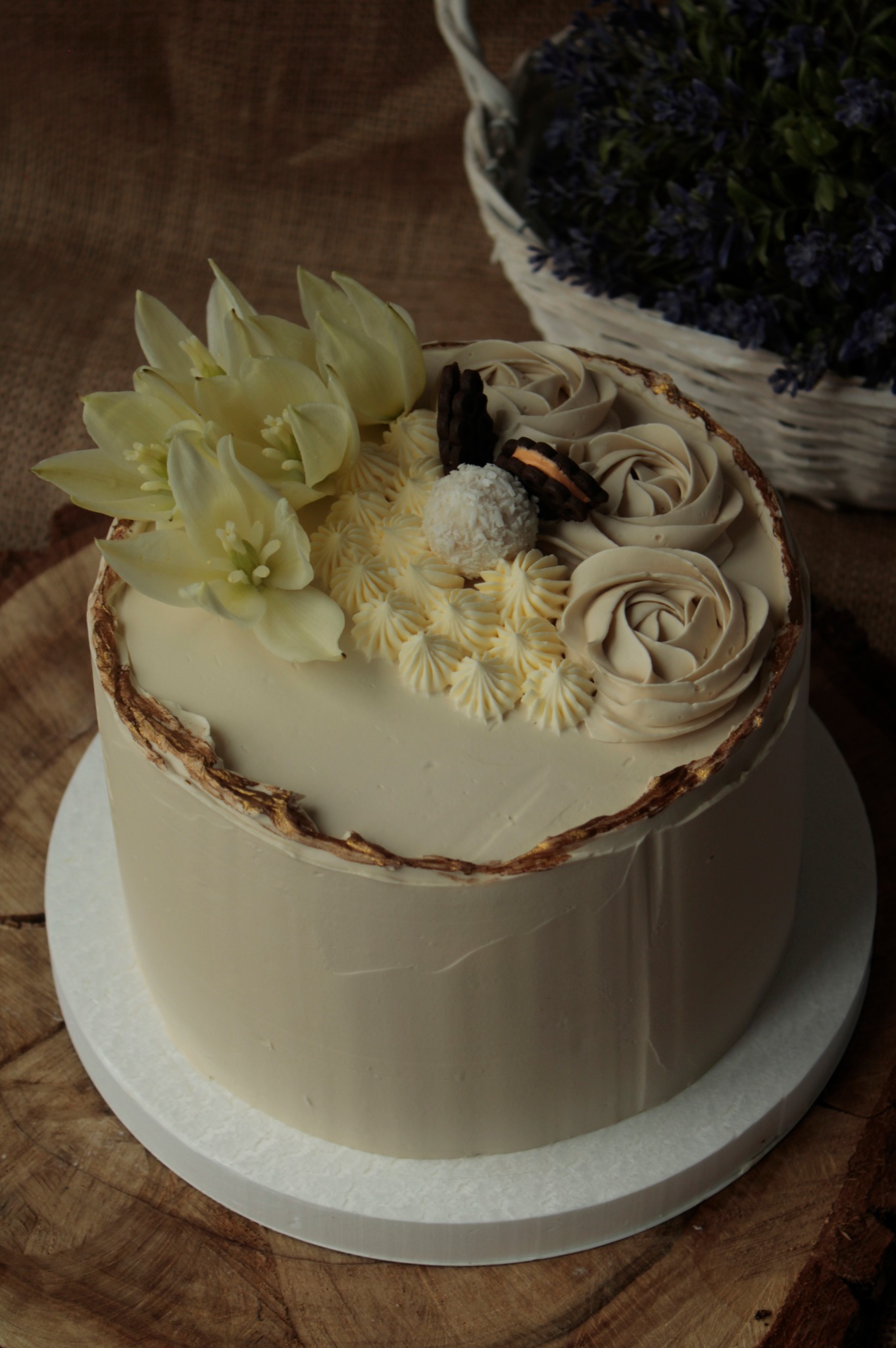 Elegancki tort z kremowym lukrem, ozdobiony białymi kwiatami lilii, rozetami z kremu i czekoladowymi ciasteczkami, wykończony złotymi akcentami na krawędzi, umieszczony na drewnianym podłożu.