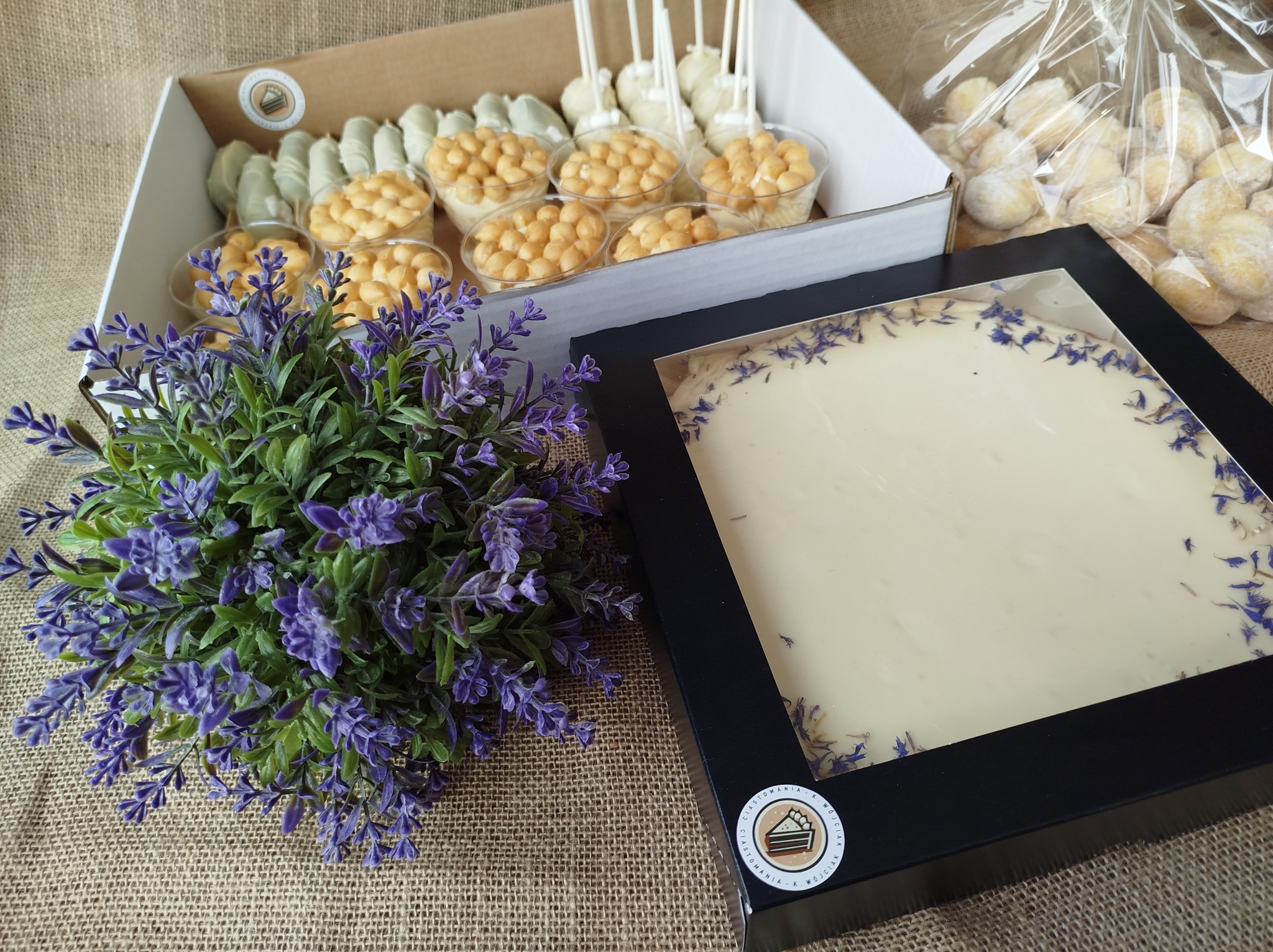 Kompozycja słodkości w pudełkach i folii na jutowym tle, z dekoracyjną lawendą: tort w czarnej ramce z białym kremem i niebieskimi płatkami, mini desery w plastikowych kubeczkach i cake pops...