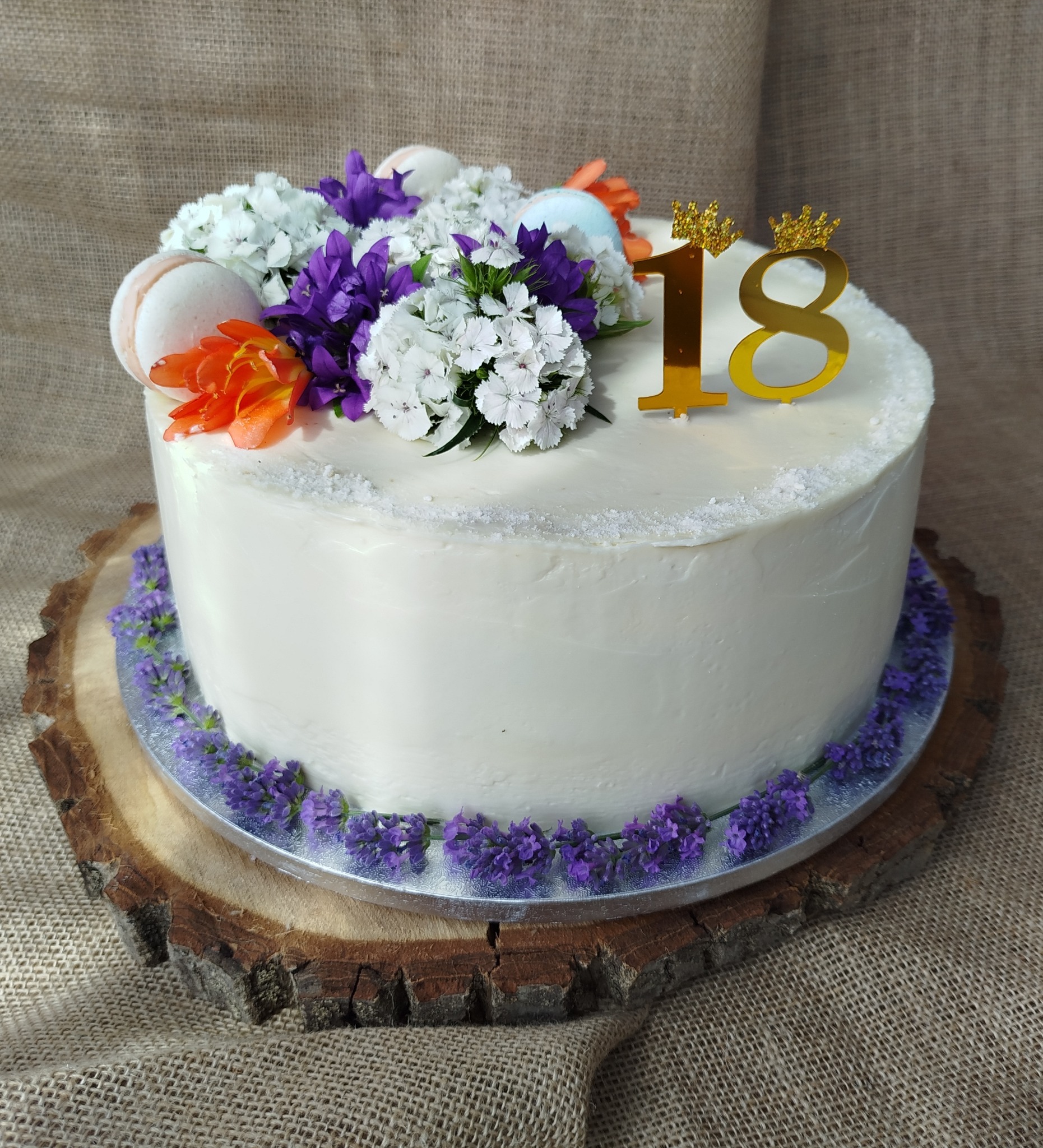 Tort urodzinowy z białym kremem, dekorowany świeżymi kwiatami w odcieniach fioletu, pomarańczy i bieli oraz makaronikami, z topperem '18' z koroną, umieszczony na drewnianej podstawie na tle...