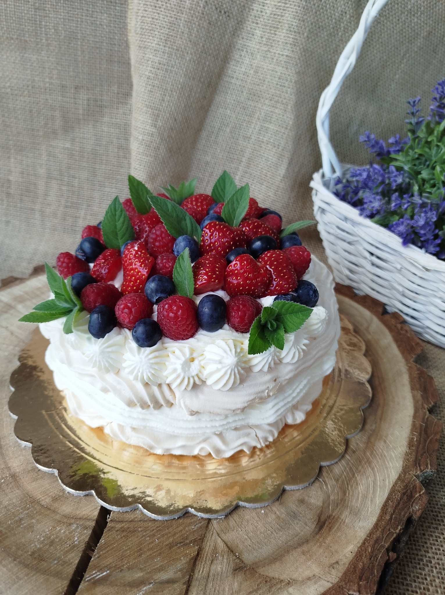 Tort bezowy udekorowany świeżymi truskawkami, malinami, borówkami i listkami mięty, na złotym podkładzie, umieszczony na drewnianym krążku z widocznymi słojami, w tle koszyk z lawendą i jutowa...