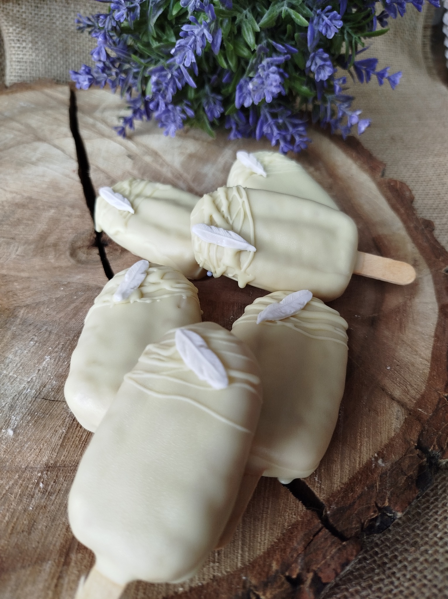 Pięć cakesicles oblanych białą czekoladą, udekorowanych białymi piórkami, ułożonych na drewnianym plastrze z pęknięciem, z dekoracją z lawendy w tle.