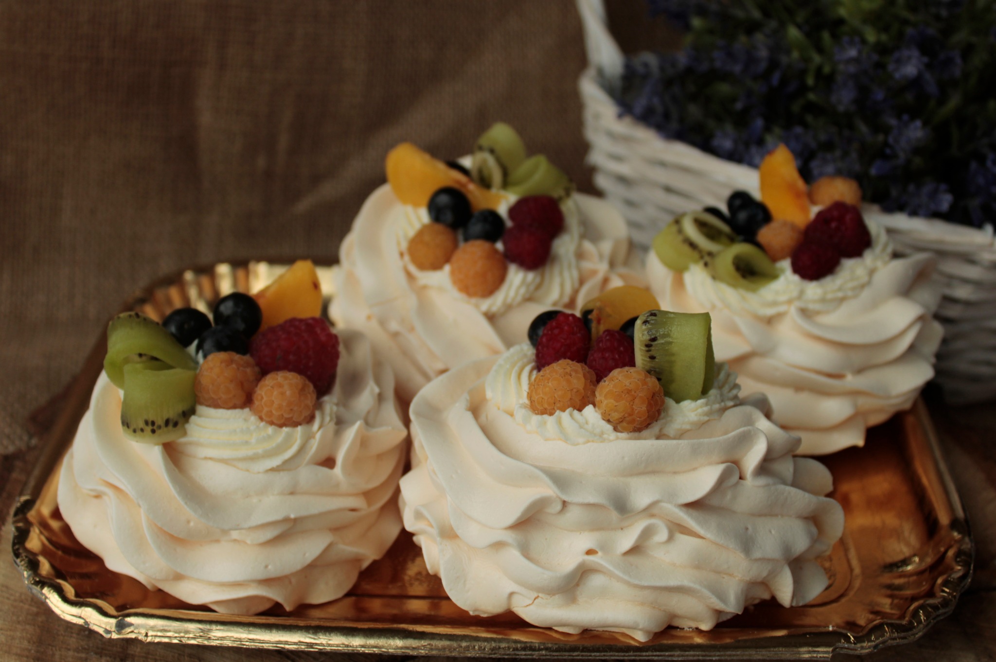 Cztery mini bezy udekorowane bitą śmietaną, malinami, borówkami, kiwi i brzoskwiniami, ułożone na złotej tacy z koszykiem lawendy w tle.