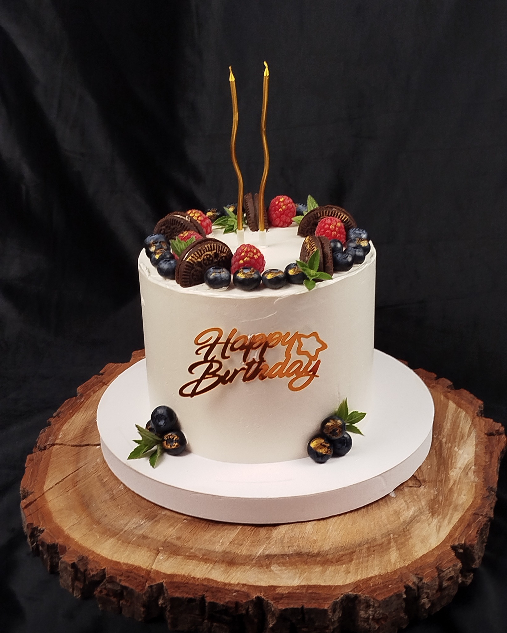 Tort urodzinowy z białym kremem, dekorowany borówkami, malinami, ciasteczkami i złotymi świeczkami, umieszczony na drewnianym plastrze. Napis 'Happy Birthday' w złotym kolorze.