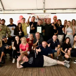 Grupowe zdjęcie zespołu firmy Raj-Pol, świętującego 25-lecie istnienia, z balonami w kształcie cyfr 25, trzymających kciuki w górę, z banerem firmy i flagą reklamową w tle, na drewnianej podłodze.