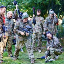 FORT BELWEDER EVENTS SPÓŁKA Z OGRANICZONĄ ODPOWIEDZIALNOŚCIĄ - Grupa uśmiechniętych osób w strojach moro grających w paintball na trawiastym terenie, niektóre osoby mają maski ochronne na twarzach, a inne trzymają markery paintballowe.