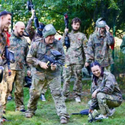 Grupa uśmiechniętych osób w strojach moro grających w paintball na trawiastym terenie, niektóre osoby mają maski ochronne na twarzach, a inne trzymają markery paintballowe.