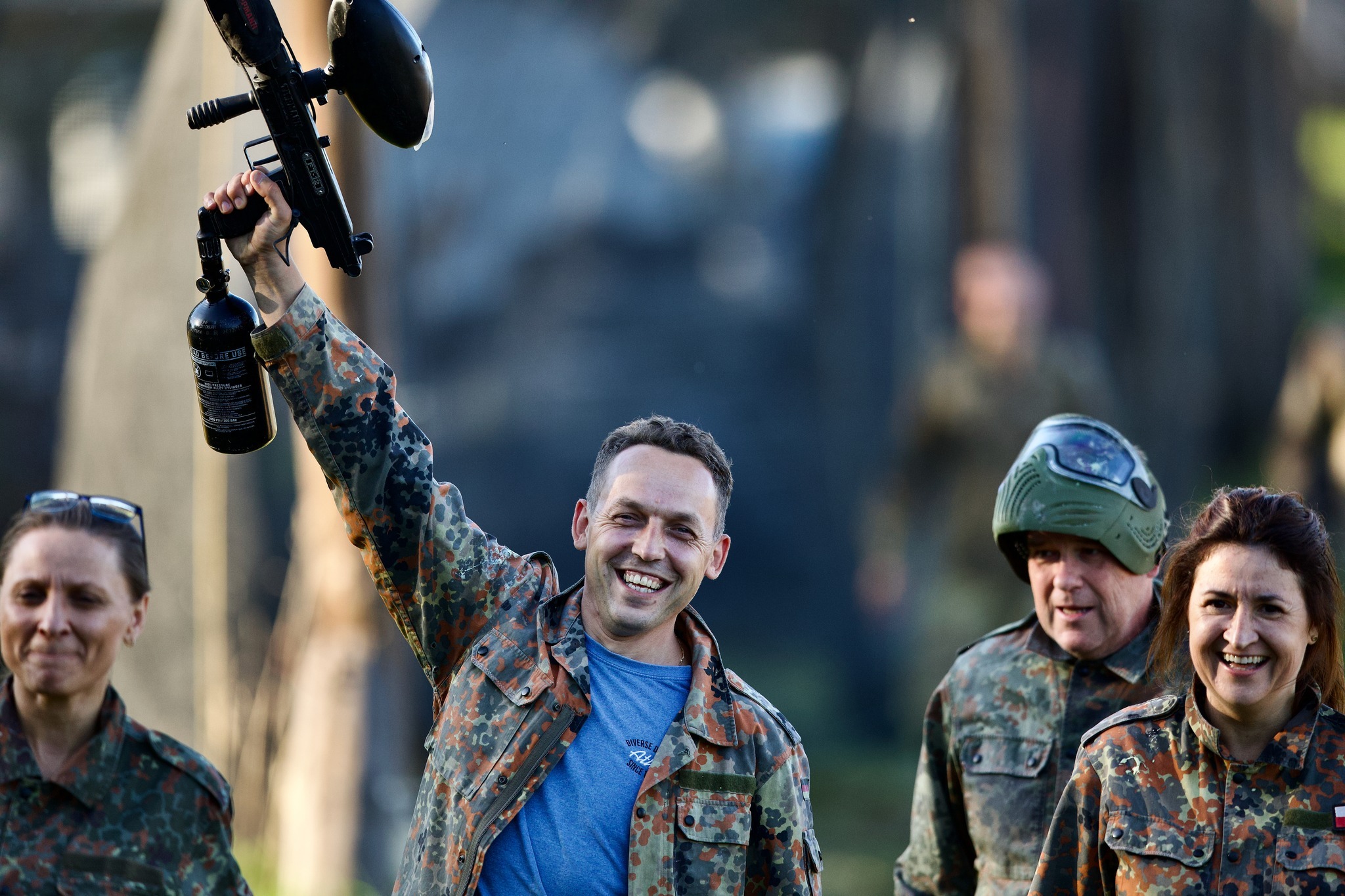 Uśmiechnięty mężczyzna w stroju moro trzyma uniesiony pistolet do paintballa z butlą CO2, na tle grupy osób w podobnych strojach i z maskami ochronnymi.