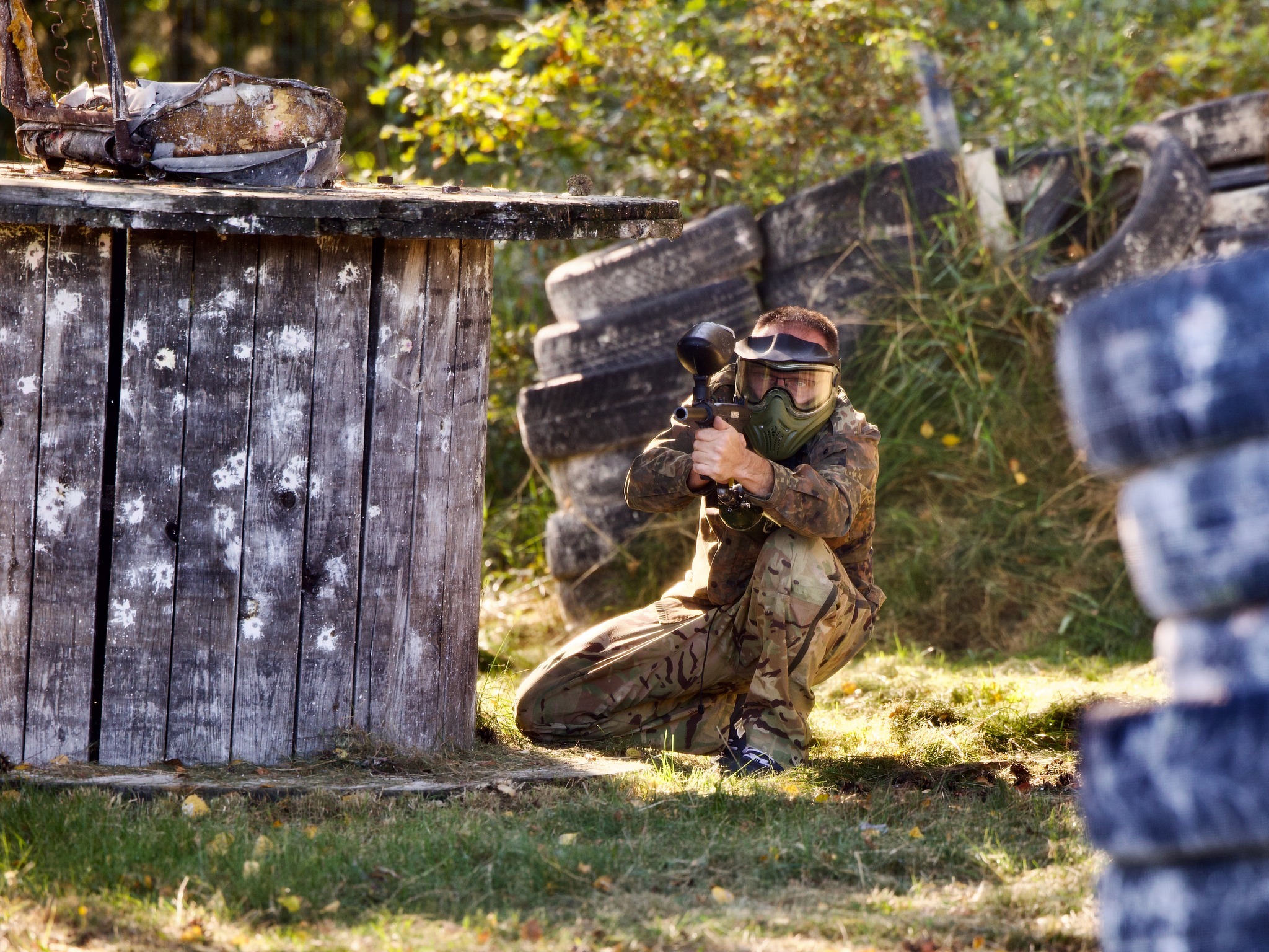 Gracz w paintballu w kamuflażu, kuca za drewnianą barykadą z widocznymi śladami farby, celując markerem w stronę widza, w tle opony i zarośla.