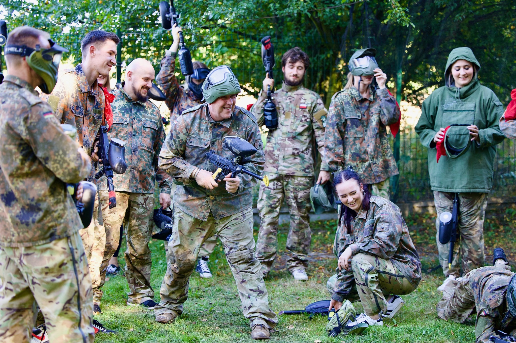 Grupa uśmiechniętych osób w strojach moro grających w paintball na trawiastym terenie, niektóre osoby mają maski ochronne na twarzach, a inne trzymają markery paintballowe.