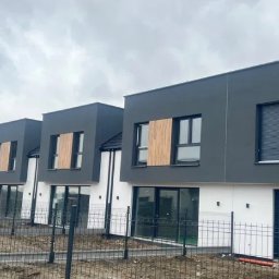 Nowoczesne, dwukondygnacyjne domy w zabudowie szeregowej z szarą elewacją i drewnianymi akcentami na oknach, ogrodzone metalowym płotem na tle pochmurnego nieba.
