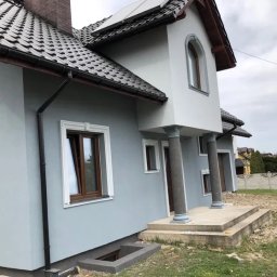 Elewacja domu jednorodzinnego w odcieniach szarości z kolumnami przy wejściu i ozdobnymi ramami okiennymi. Widoczne detale wykończenia i dach z ciemnej dachówki.