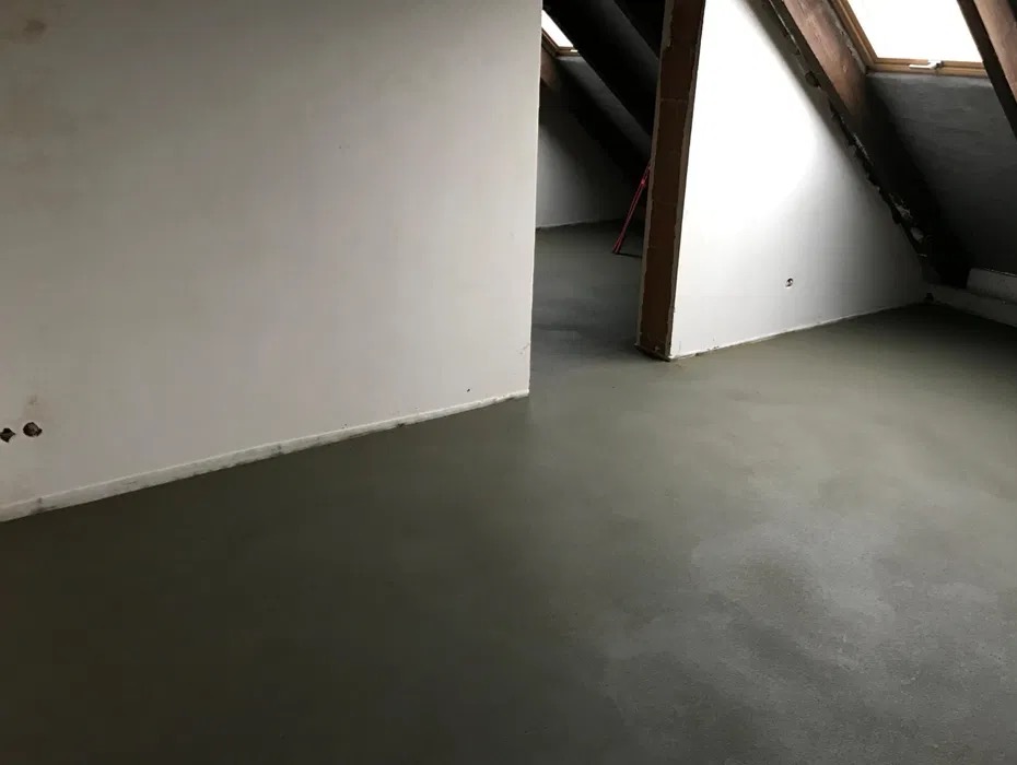 Świeżo wylana, gładka posadzka jastrychowa w jasnym pomieszczeniu na poddaszu z oknami dachowymi i drewnianą konstrukcją więźby.