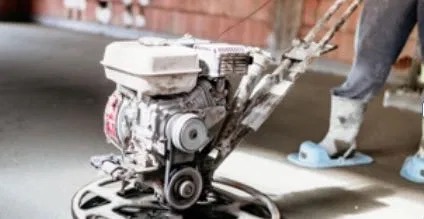 Zacieraczka mechaniczna w trakcie pracy na świeżo wylanej posadzce jastrychowej, widoczne buty ochronne pracownika i fragment muru z cegły.