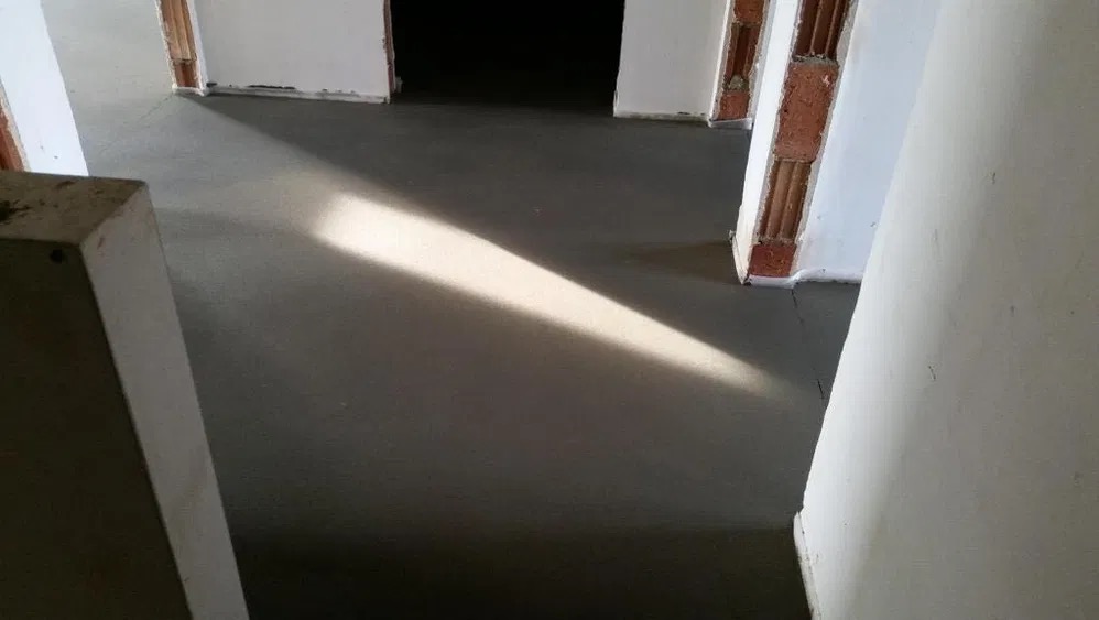 Świeżo wylana, gładka posadzka w jasnym pomieszczeniu z surowymi, ceglanymi elementami ścian i wyraźnym pasmem światła słonecznego padającym na powierzchnię.