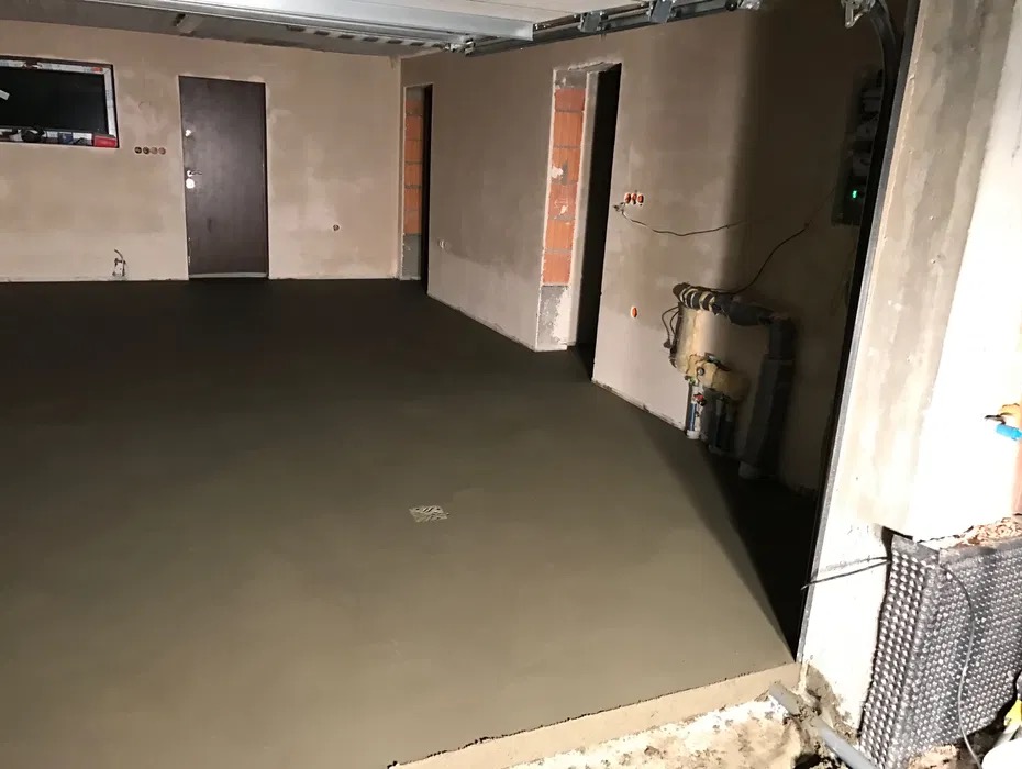 Świeżo wylana, gładka posadzka jastrychowa w surowym wnętrzu, widoczne instalacje na ścianie i otwór drzwiowy z ceglanym obramowaniem.