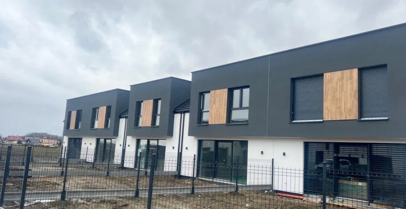 Nowoczesne, dwukondygnacyjne domy w zabudowie szeregowej z szarą elewacją i drewnianymi akcentami na oknach, ogrodzone metalowym płotem na tle pochmurnego nieba.