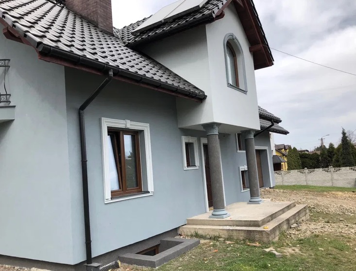Elewacja domu jednorodzinnego w odcieniach szarości z kolumnami przy wejściu i ozdobnymi ramami okiennymi. Widoczne detale wykończenia i dach z ciemnej dachówki.