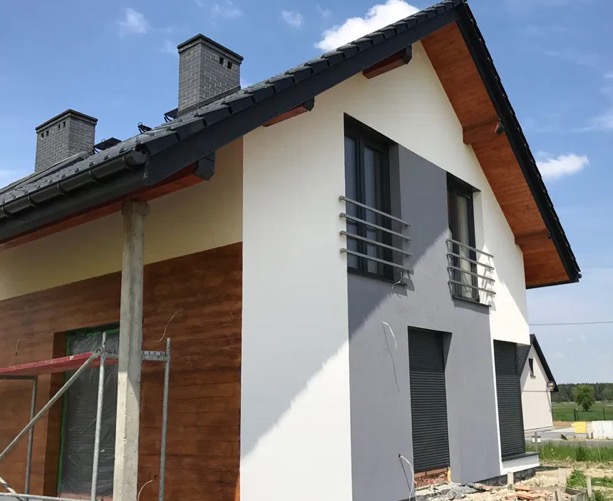 Dwukondygnacyjny dom z elewacją w odcieniach bieli i szarości, z elementami dekoracyjnymi imitującymi drewno. Widoczne rusztowanie sugeruje trwające prace wykończeniowe.