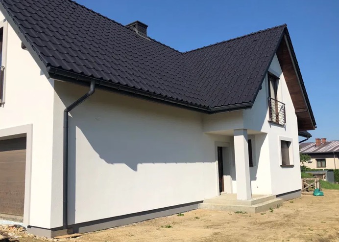 Biały dom jednorodzinny z ciemną dachówką i szarymi elementami wykończeniowymi, widoczny z zewnątrz w słoneczny dzień. Elewacja świeżo po remoncie.