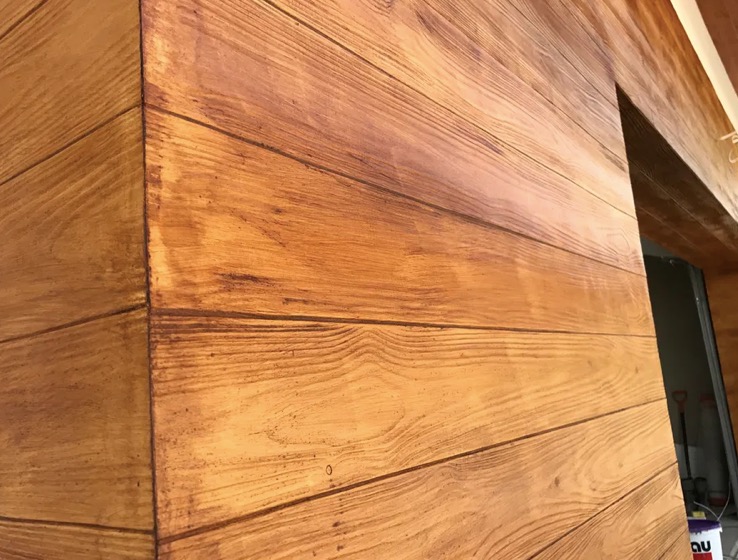 Ściana wewnętrzna wykończona w stylu drewnianych paneli, z widoczną fakturą i słojami drewna, pomalowana na ciepły, brązowy odcień. Widoczny fragment futryny drzwi.