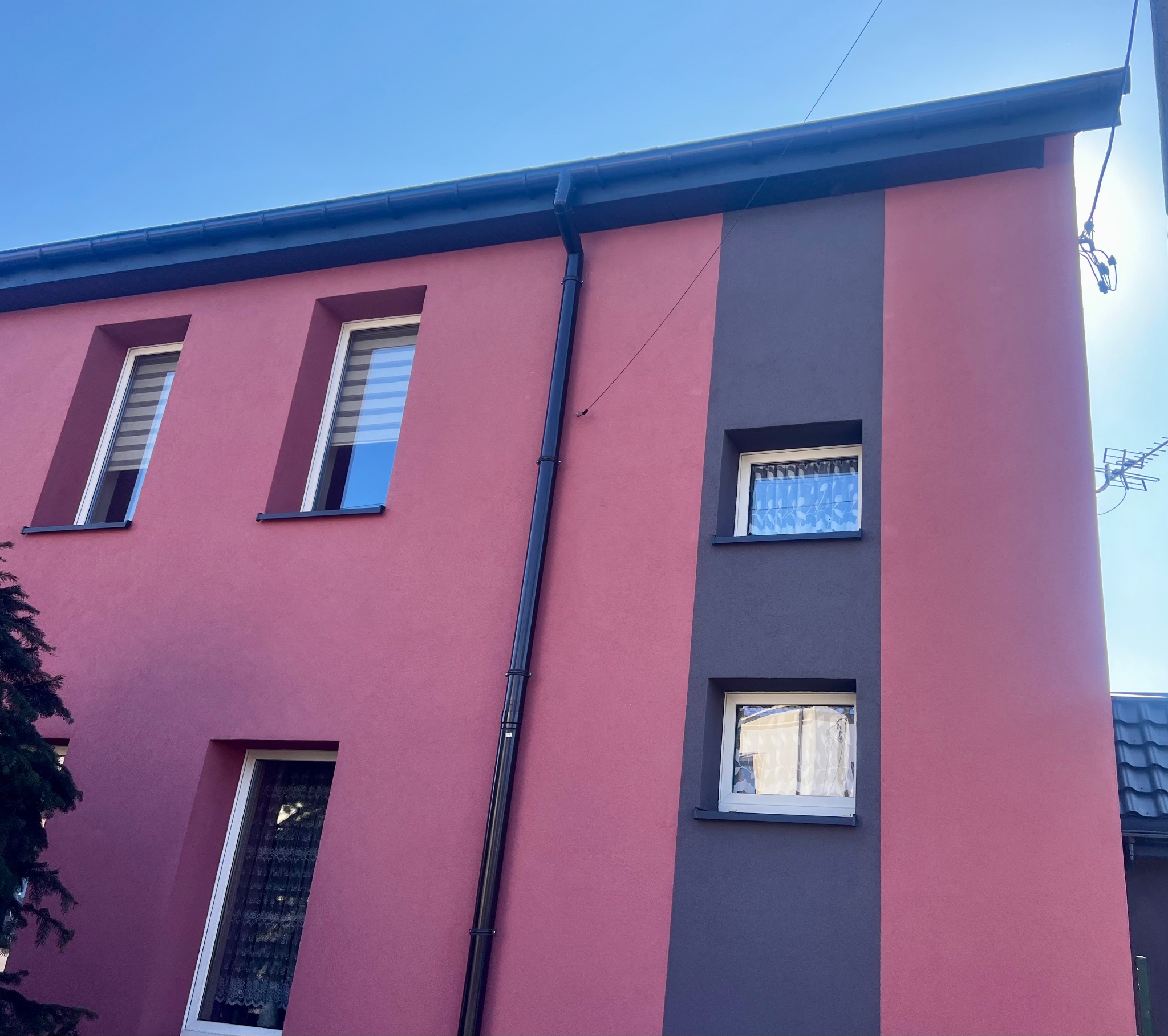 Dwukolorowa elewacja domu z oknami, rynną i anteną. Część ściany w odcieniu różu, druga w kolorze grafitowym. Widok z dołu, błękitne niebo w tle.