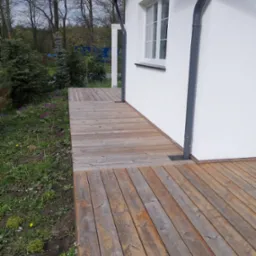 Drewniany taras przylegający do białej ściany domu z szarą rynną, widok z poziomu gruntu, zieleń i drzewa w tle.