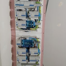 Artur Mroziński - Dwie rozdzielnice elektryczne zamontowane na ścianie z widocznymi bezpiecznikami, przewodami w kolorach niebieskim, zielonym i żółtym oraz modułami zabezpieczającymi. Widoczne oznaczenia...