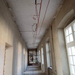 Artur Mroziński - Korytarz w trakcie remontu z widocznymi przewodami elektrycznymi prowadzonymi po suficie, drabina w tle i osoba w oddali.