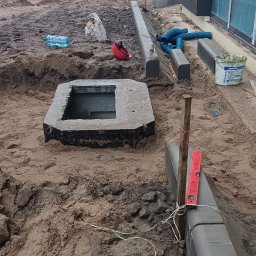 Artur Mroziński - Przygotowanie terenu pod instalację elektryczną zewnętrzną: wykopana studzienka z betonową obudową, obok ułożone krawężniki, poziomica, rury osłonowe i butelka wody na placu budowy.