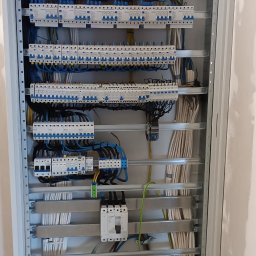 Artur Mroziński - Rozdzielnica elektryczna w metalowej obudowie, widoczne liczne zabezpieczenia nadprądowe, przewody w kolorach niebieskim, żółto-zielonym i brązowym, ułożone wewnątrz szafy. Część przewodów...