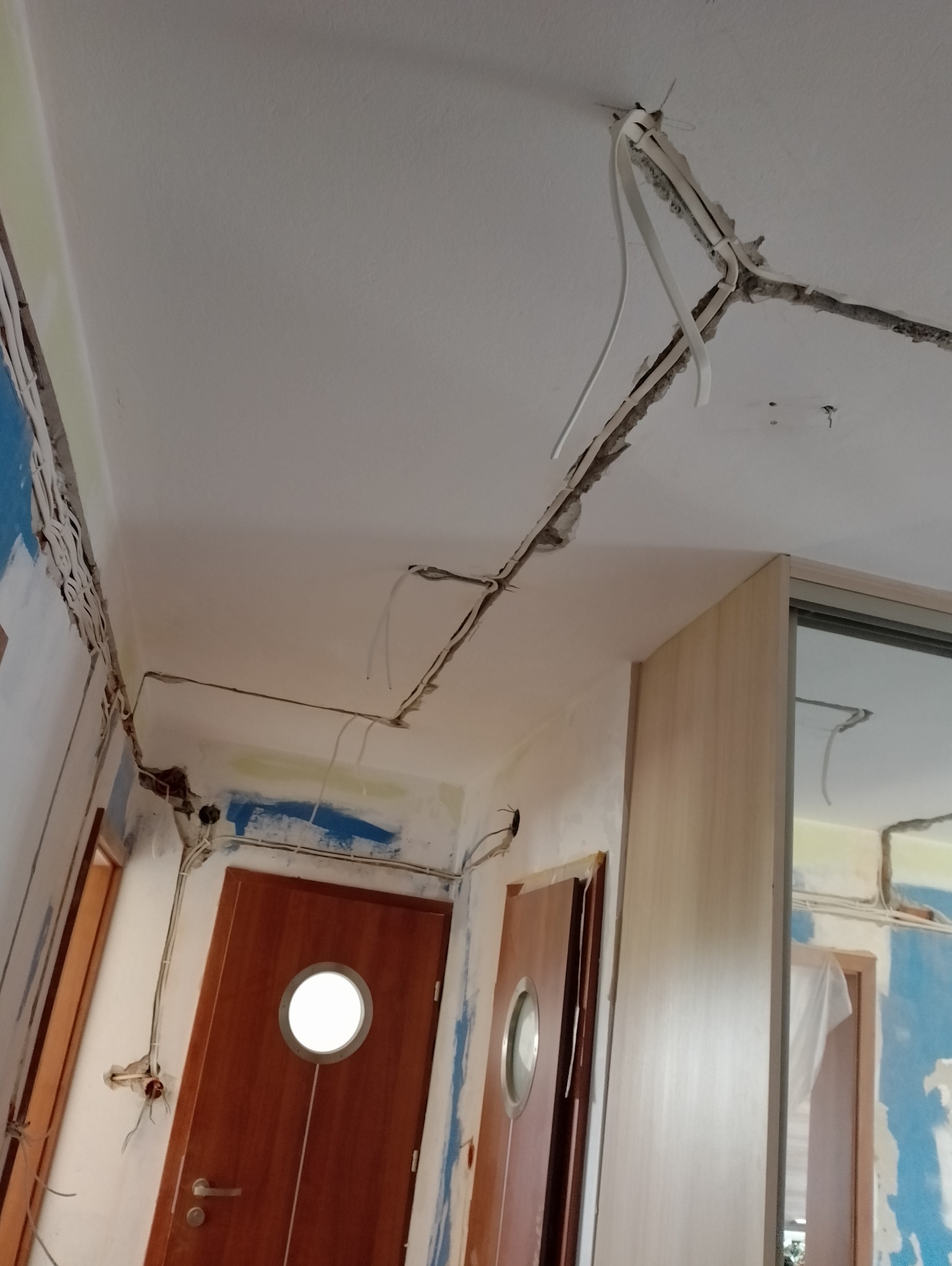 Wnętrze w trakcie remontu z widocznymi przewodami elektrycznymi ułożonymi w bruzdach na suficie i ścianach, przygotowanymi do zalania gipsem. Na ścianach i suficie widoczne ślady po kuciu. W tle...