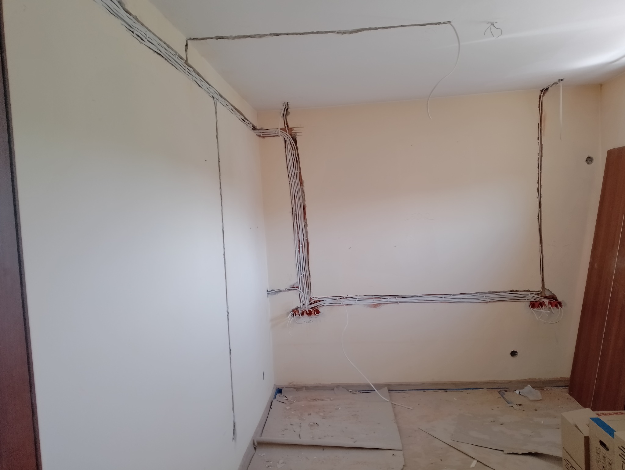 Wnętrze pokoju w trakcie remontu z widocznymi przewodami elektrycznymi ułożonymi w bruzdach ściennych i pod sufitem, puszki elektryczne zamontowane w ścianie, podłoga zabezpieczona kartonem.