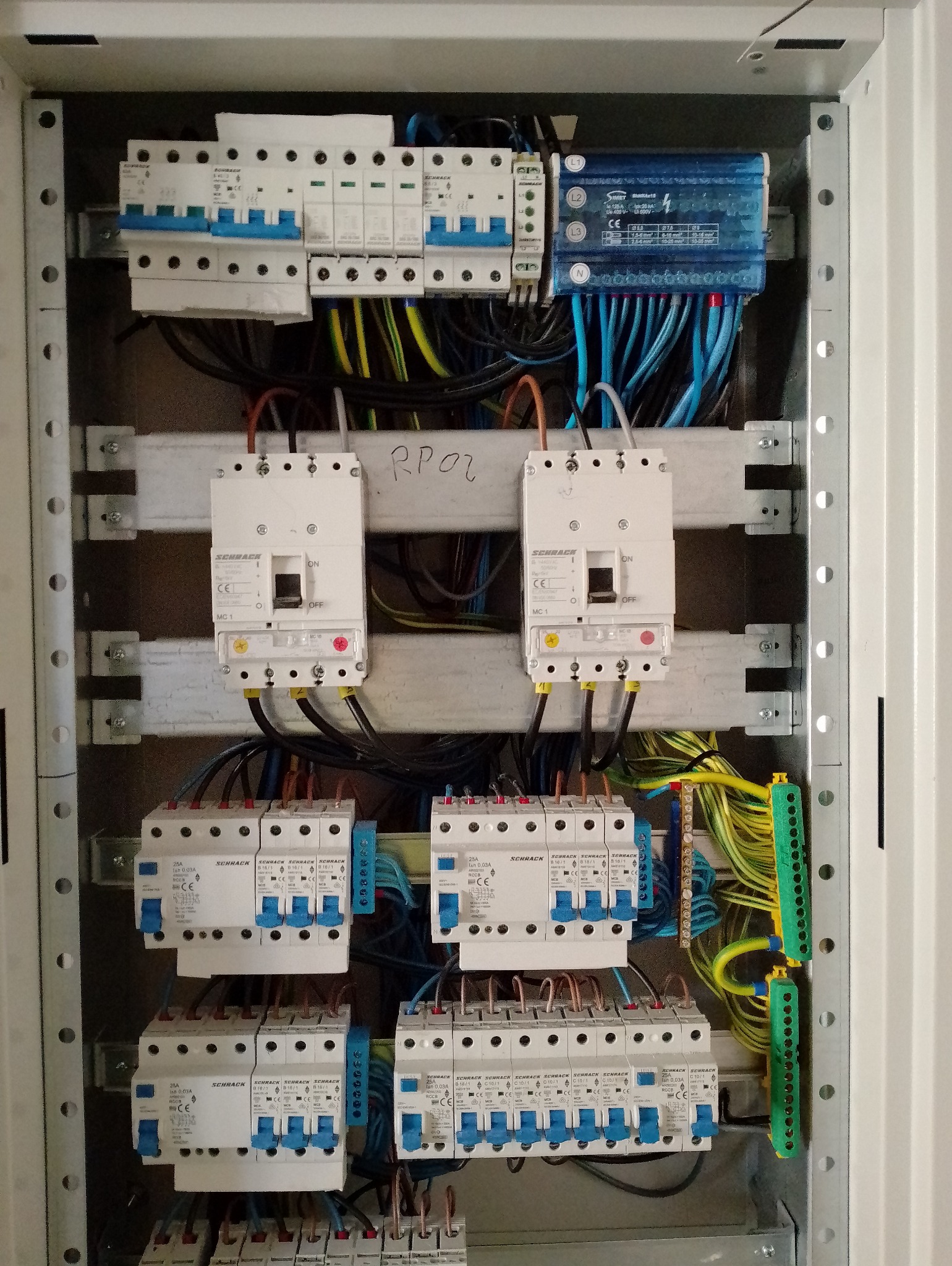 Rozdzielnica elektryczna z zainstalowanymi bezpiecznikami Schrack, zabezpieczeniami różnicowoprądowymi i listwami zaciskowymi, widoczne przewody w różnych kolorach, opis RPO2 na szynie.
