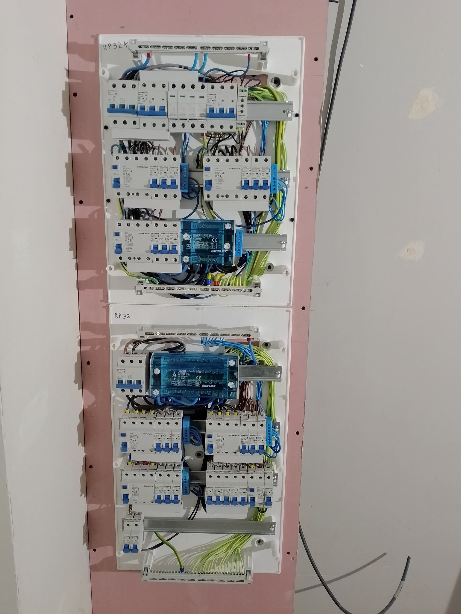 Dwie rozdzielnice elektryczne zamontowane na ścianie z widocznymi bezpiecznikami, przewodami w kolorach niebieskim, zielonym i żółtym oraz modułami zabezpieczającymi. Widoczne oznaczenia...