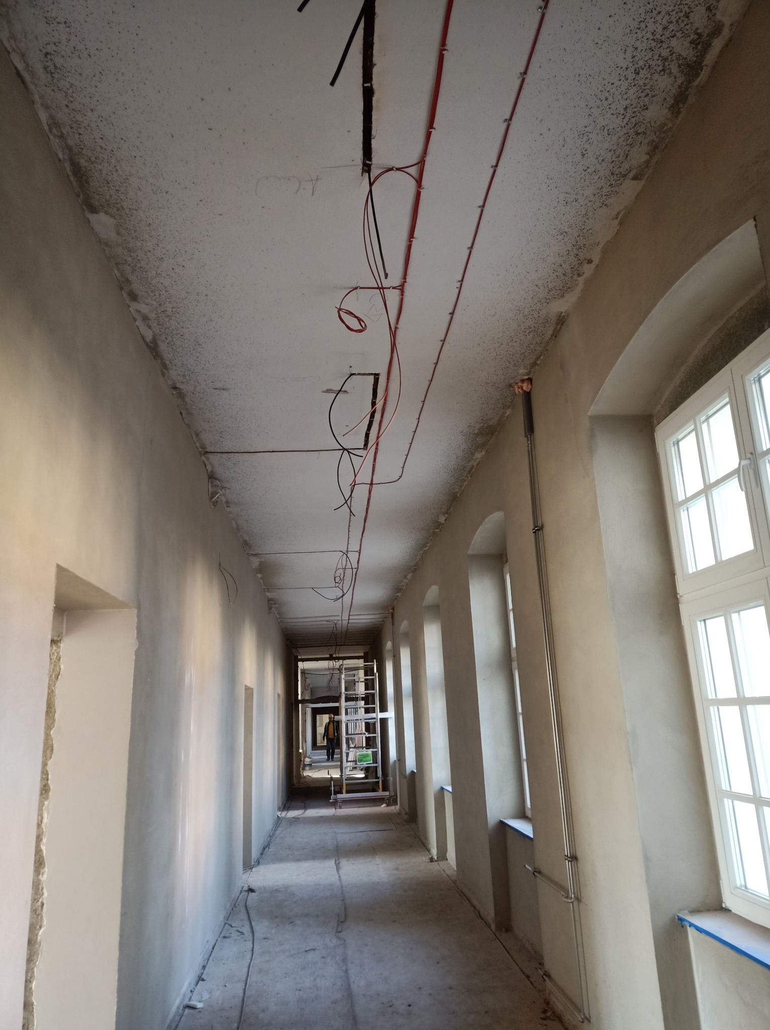Korytarz w trakcie remontu z widocznymi przewodami elektrycznymi prowadzonymi po suficie, drabina w tle i osoba w oddali.