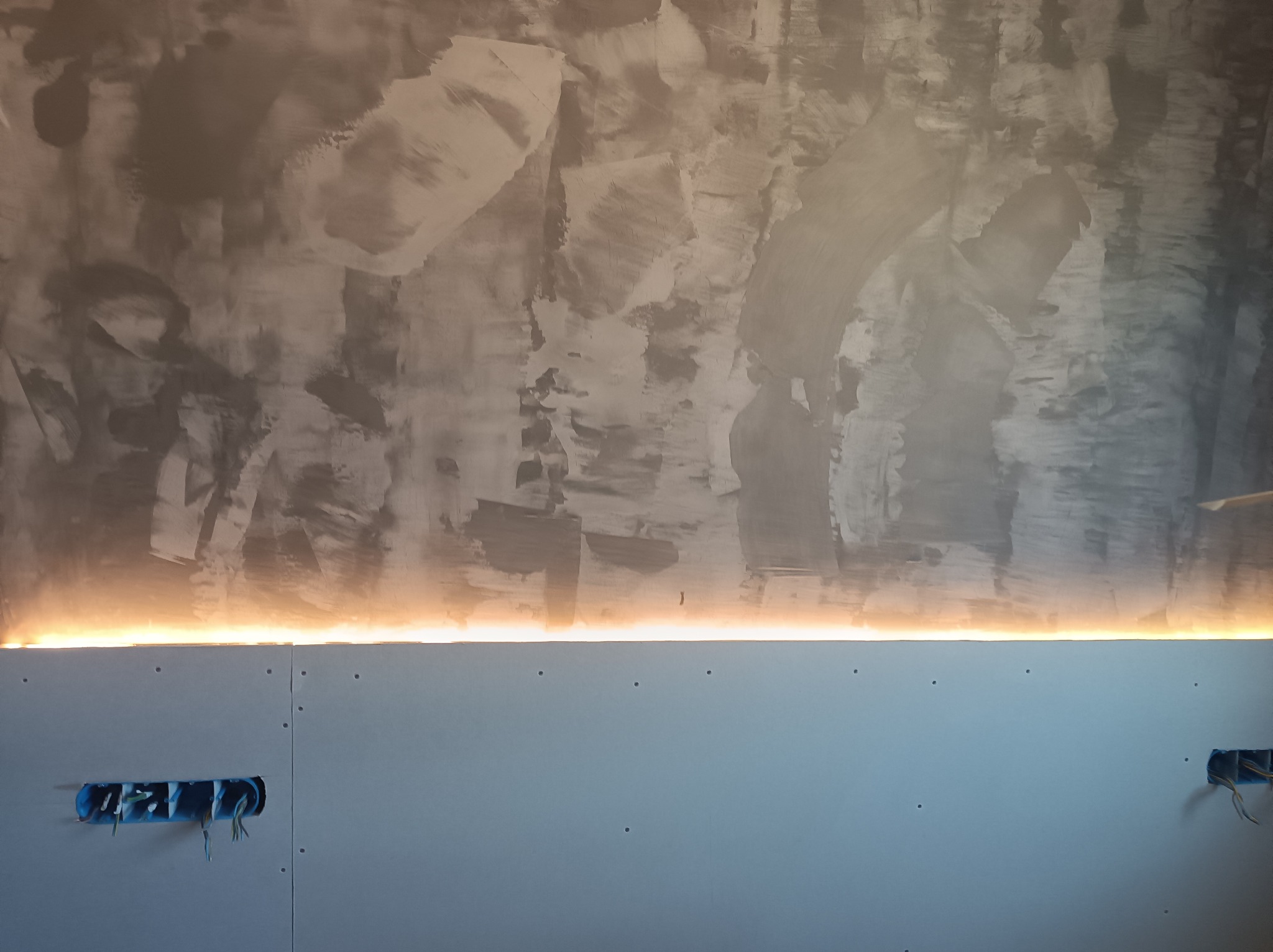 Ściana z szarą, nieregularną fakturą i zamontowanym oświetleniem LED w szczelinie, widoczne puszki elektryczne z wystającymi przewodami, płyty gipsowo-kartonowe.