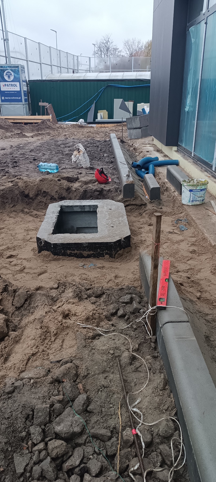 Przygotowanie terenu pod instalację elektryczną zewnętrzną: wykopana studzienka z betonową obudową, obok ułożone krawężniki, poziomica, rury osłonowe i butelka wody na placu budowy.