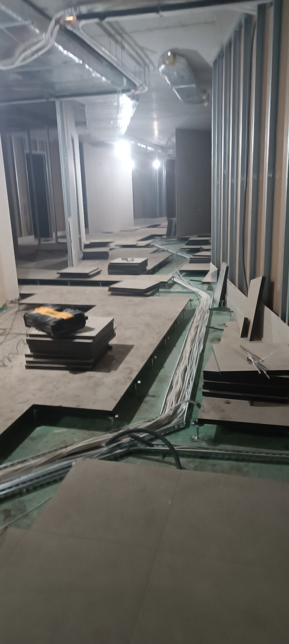 Wnętrze w trakcie instalacji elektrycznej: widoczne kable prowadzone w podłodze, metalowe profile ścian, narzędzie elektryczne na stosie płyt podłogowych.