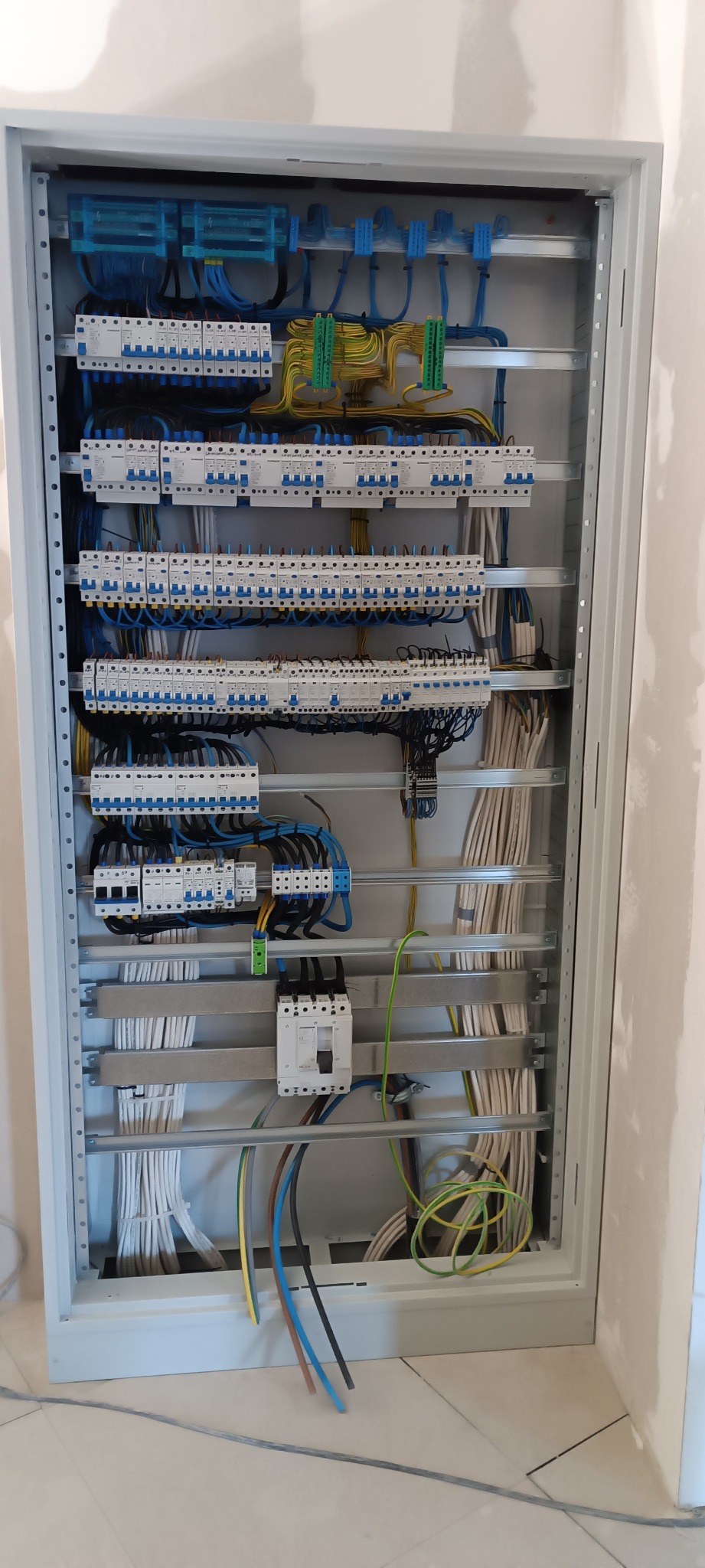 Rozdzielnica elektryczna w metalowej obudowie, widoczne liczne zabezpieczenia nadprądowe, przewody w kolorach niebieskim, żółto-zielonym i brązowym, ułożone wewnątrz szafy. Część przewodów...