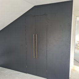 MARTENS HAUSE DANIEL BOROWSKI - Grafitowa szafa wnękowa z długimi, złotymi uchwytami, wkomponowana w skos dachu, widok z boku na surową przestrzeń w trakcie wykończenia.