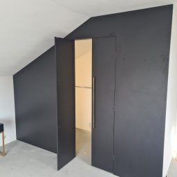 MARTENS HAUSE DANIEL BOROWSKI - Czarna szafa wnękowa z otwartymi drzwiami w skosie poddasza, widoczny drążek na ubrania i mała szafka nocna w podobnym stylu.