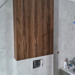 MARTENS HAUSE DANIEL BOROWSKI - Wysoka szafka łazienkowa z drewnianym frontem, zamontowana nad spłuczką w toalecie wykończonej jasnymi, wzorzystymi płytkami.