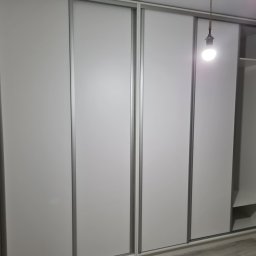 MARTENS HAUSE DANIEL BOROWSKI - Biała szafa wnękowa z przesuwnymi drzwiami i widocznymi półkami z boku, oświetlona wiszącą żarówką na kablu.