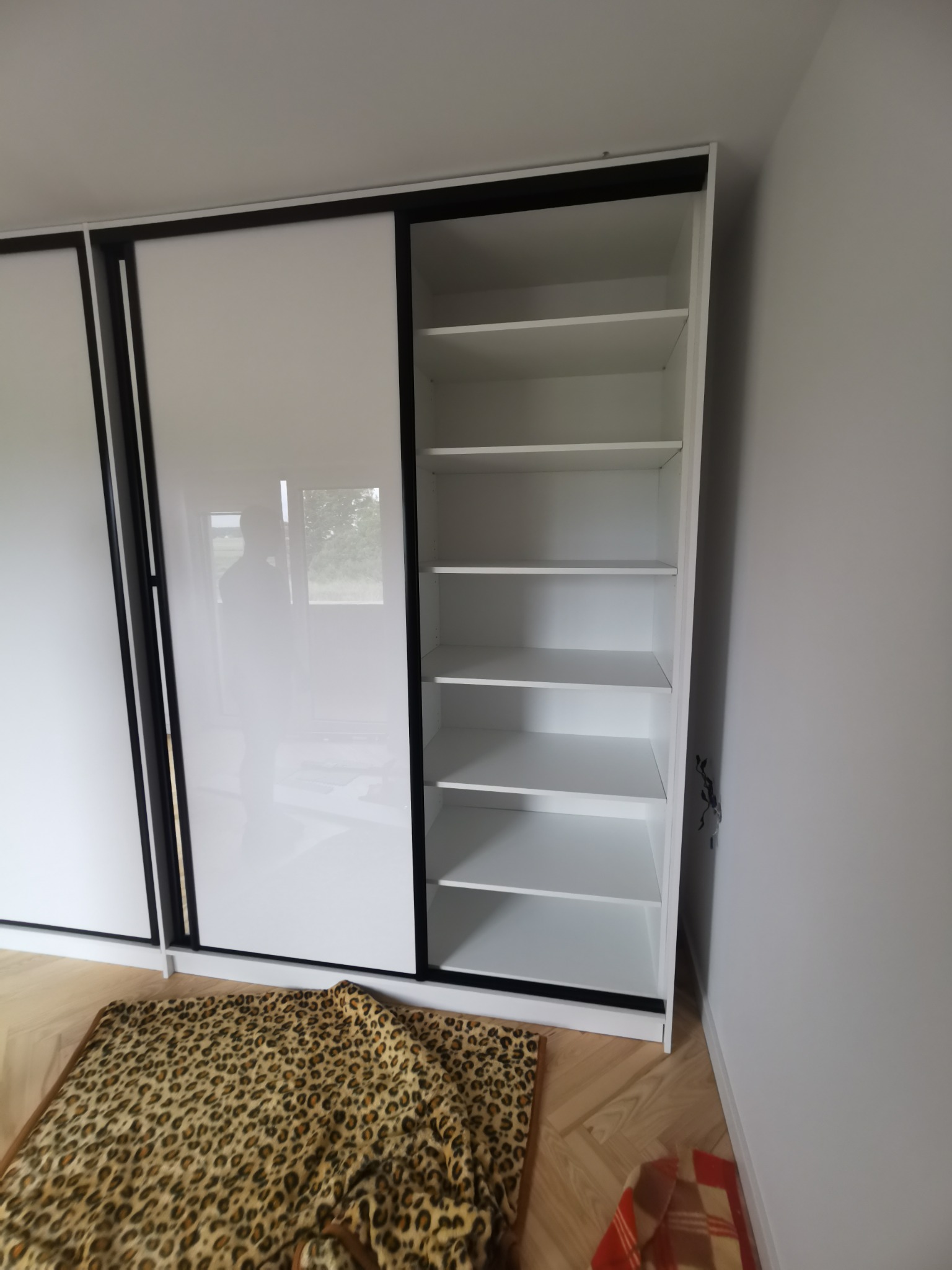 Biała szafa z przesuwnymi drzwiami i otwartymi półkami, widoczny fragment podłogi z wzorem jodełki i koc w panterkę.