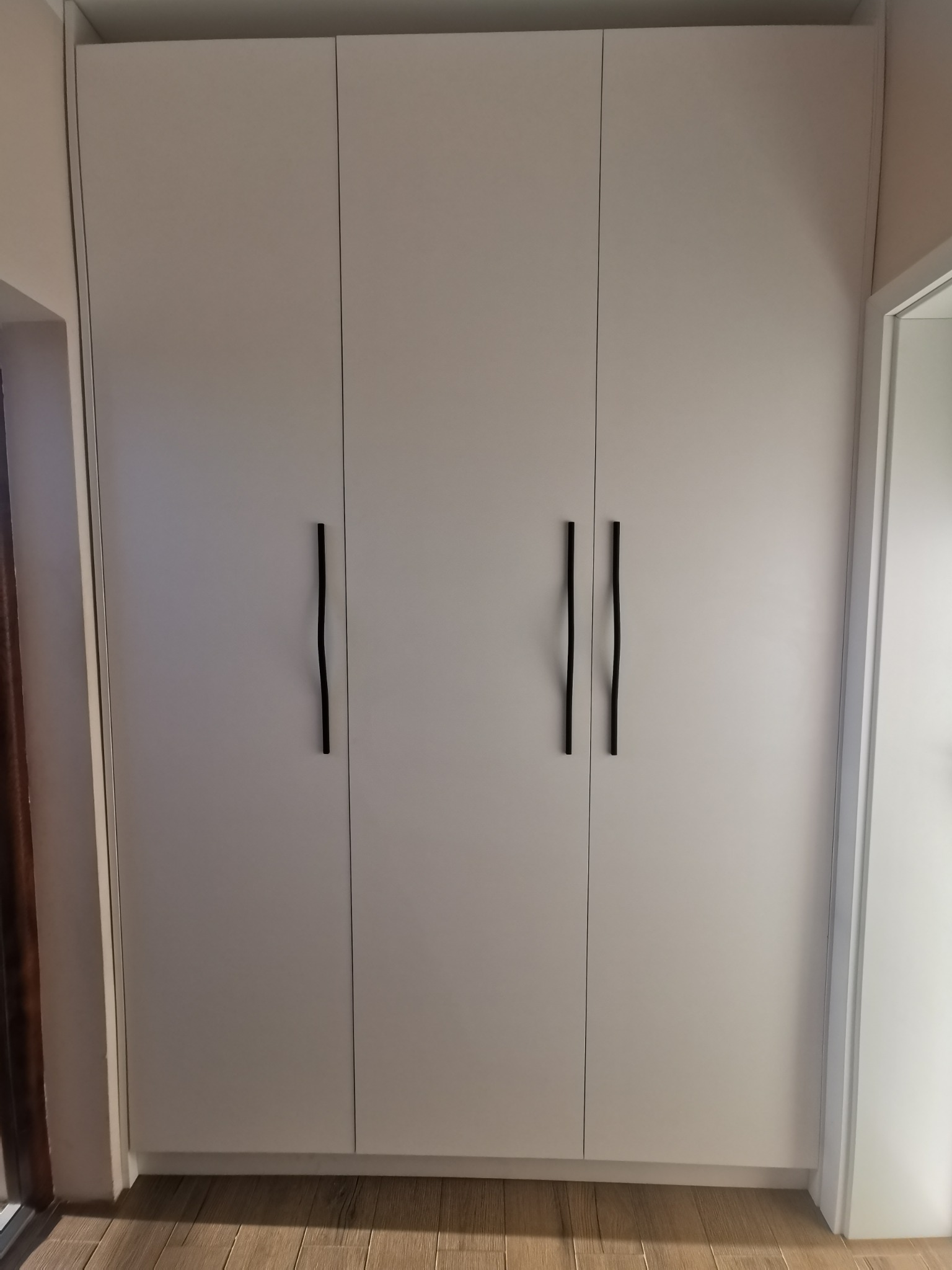 Biała, trzydrzwiowa szafa wnękowa z długimi, czarnymi uchwytami na tle drewnianej podłogi i fragmentu drzwi.