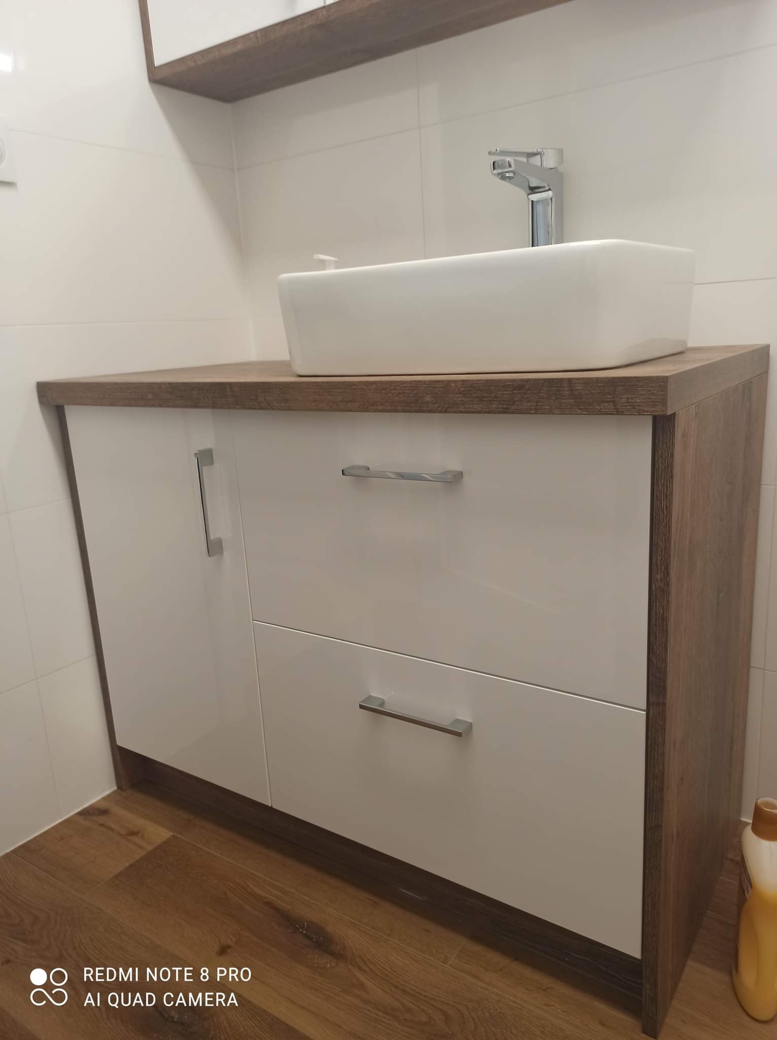 Nowoczesna biała umywalka nablatowa na szafce z białymi frontami i drewnianym blatem w łazience z jasnymi płytkami.