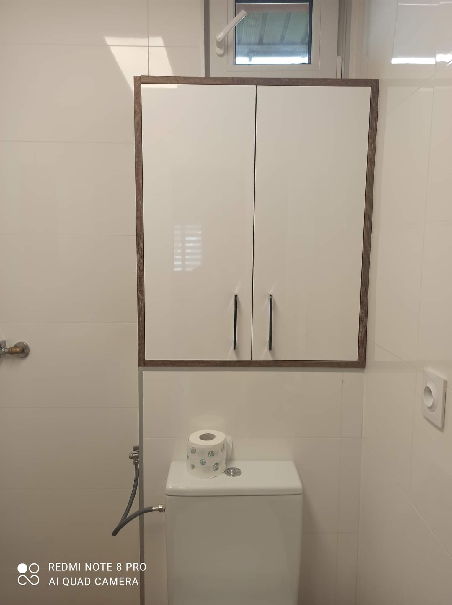 Biała szafka wisząca z drewnianą ramą nad toaletą w wykończonej łazience z białymi płytkami.