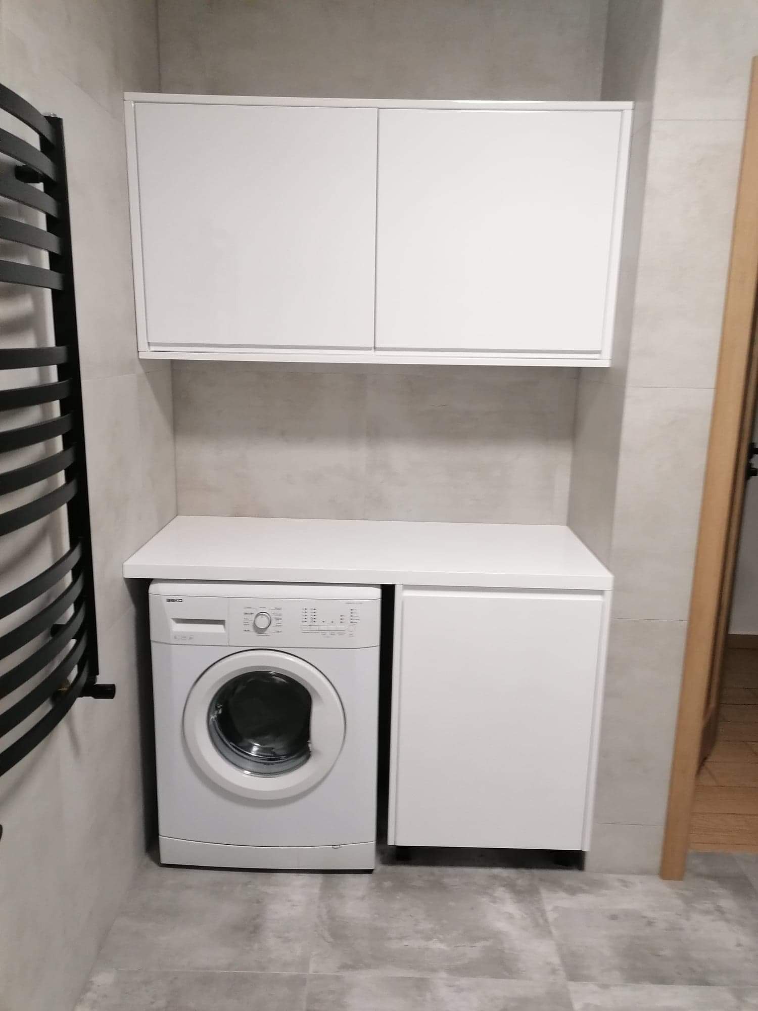 Biała pralka zabudowana pod blatem z szafką i wiszącą szafką, szary płytki na ścianach i podłodze, czarny grzejnik drabinkowy z boku.